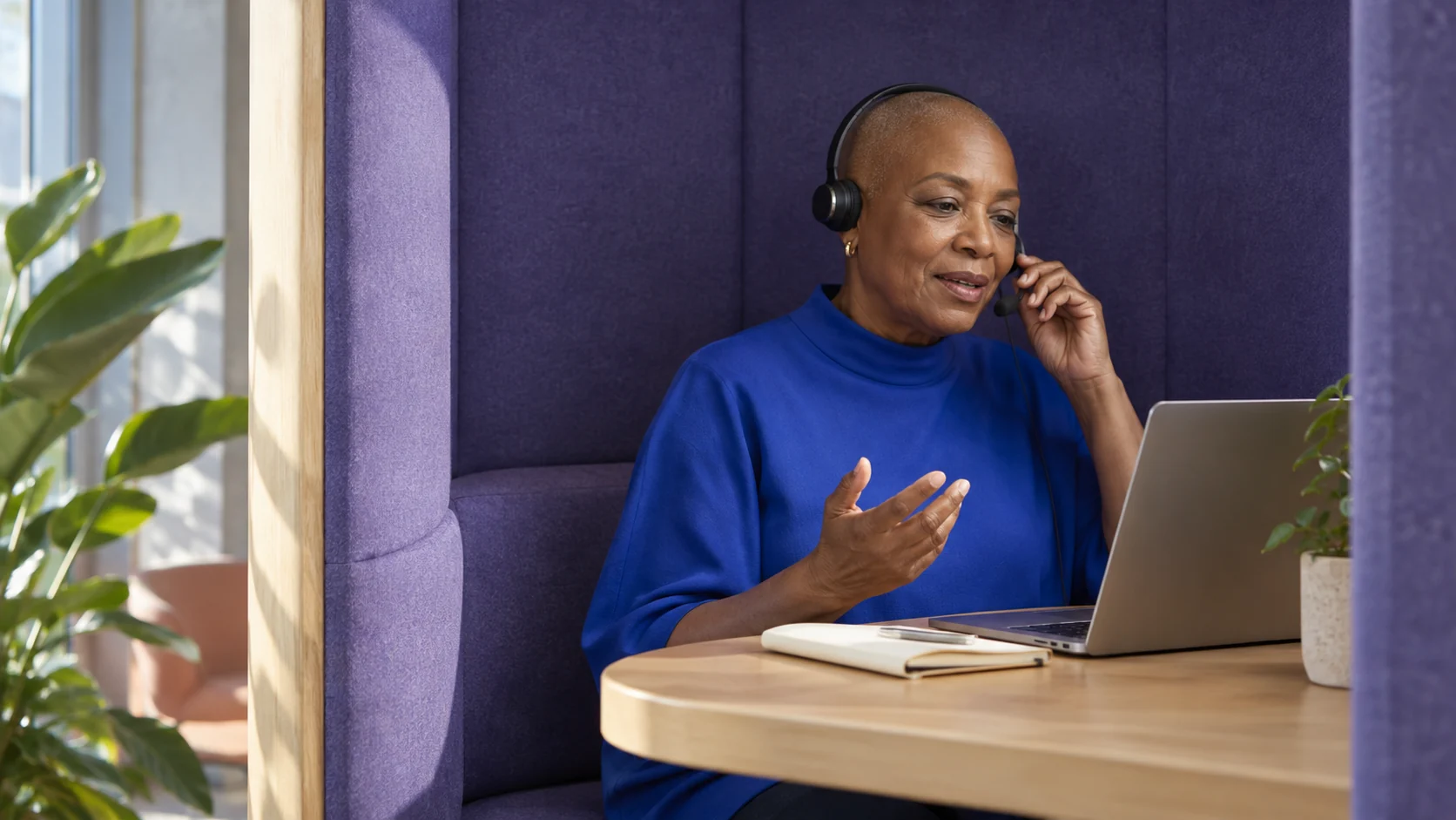 A woman wearing a headset speaks from a violet acoustic booth with a notebook and laptop on its pale wood ledge.